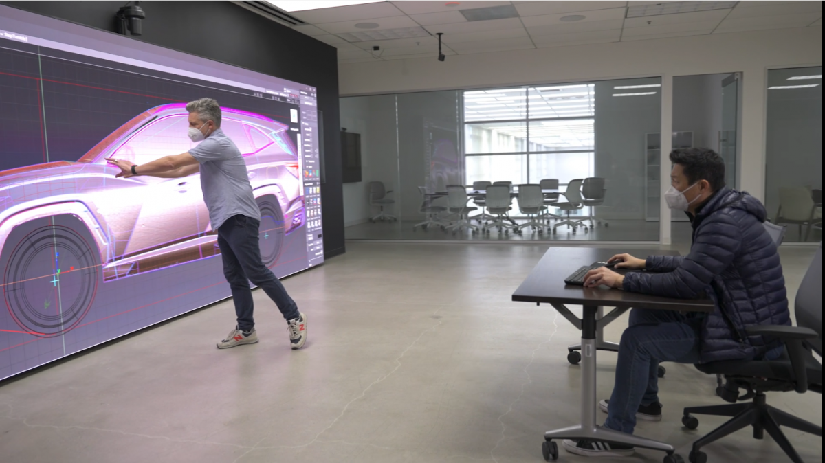 Employee in front of a large screen displaying a car design schematic in front of a coworker sitting at a desk