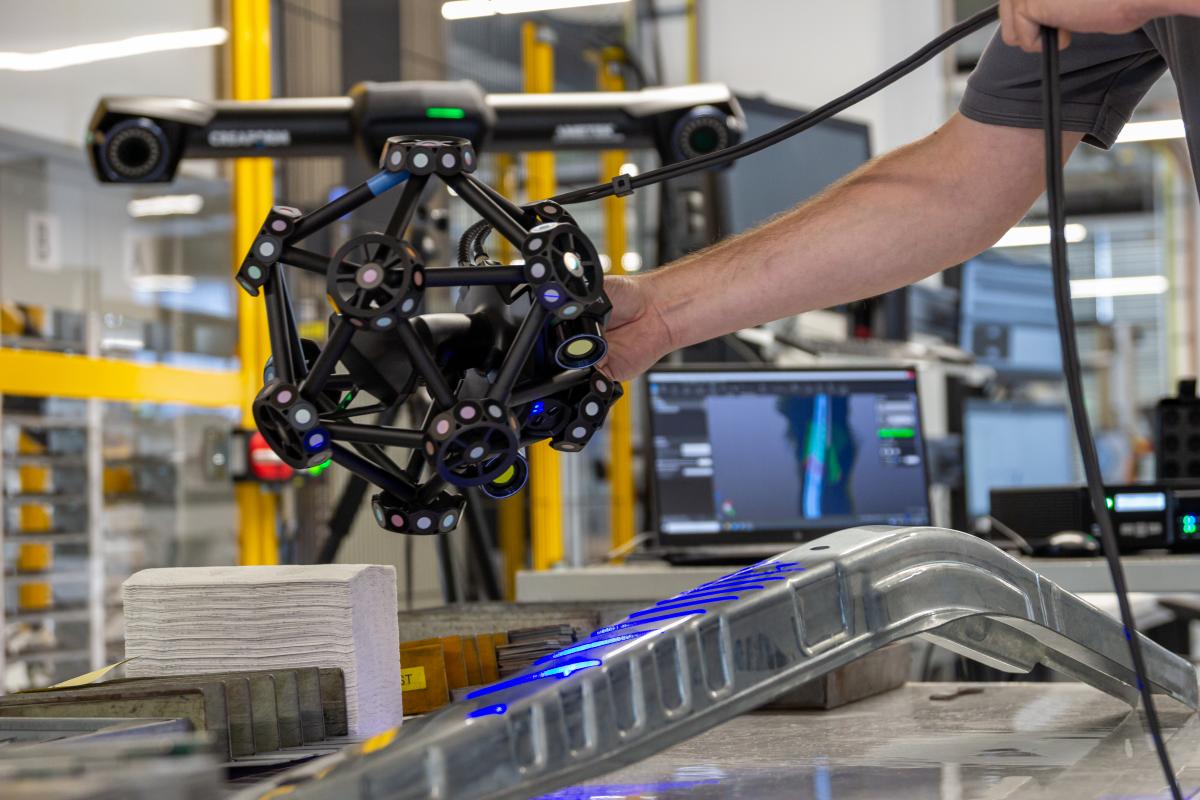 El escáner MetraSCAN 3D que escanea una pieza de chapa. En segundo plano, puede ver la grabación en tiempo real del escaneado en la computadora portátil.