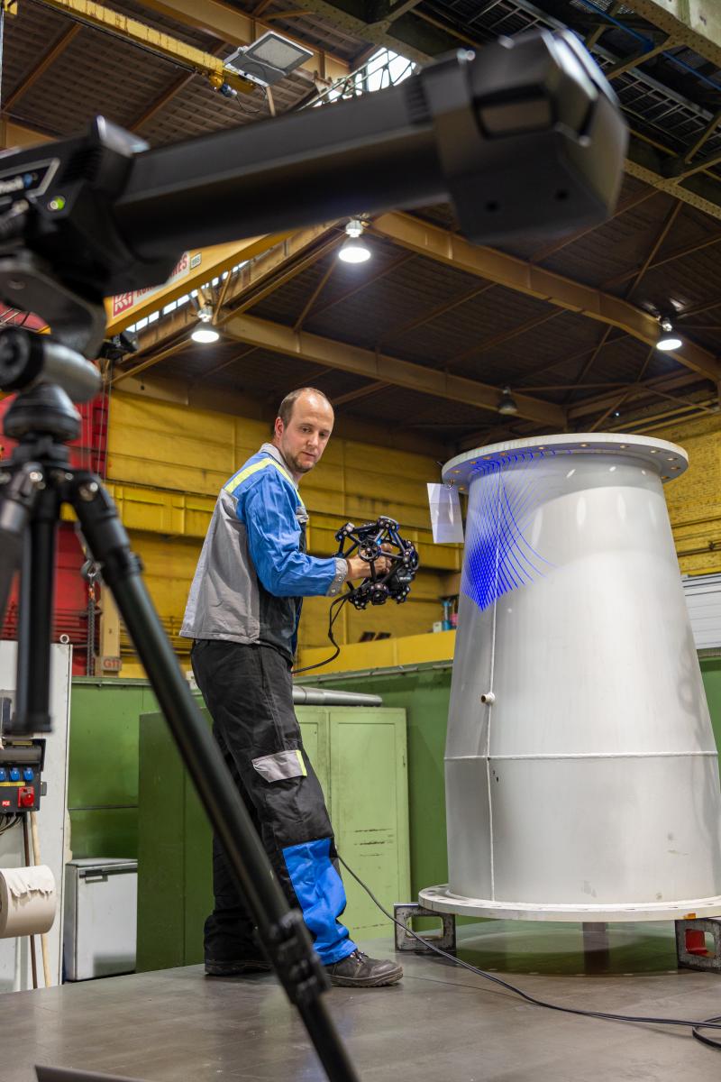 Un hombre escanea un segmento de tubo de un alto horno con el MetraSCAN 3D. En primer plano está la barra de la cámara C-Track sobre un trípode.