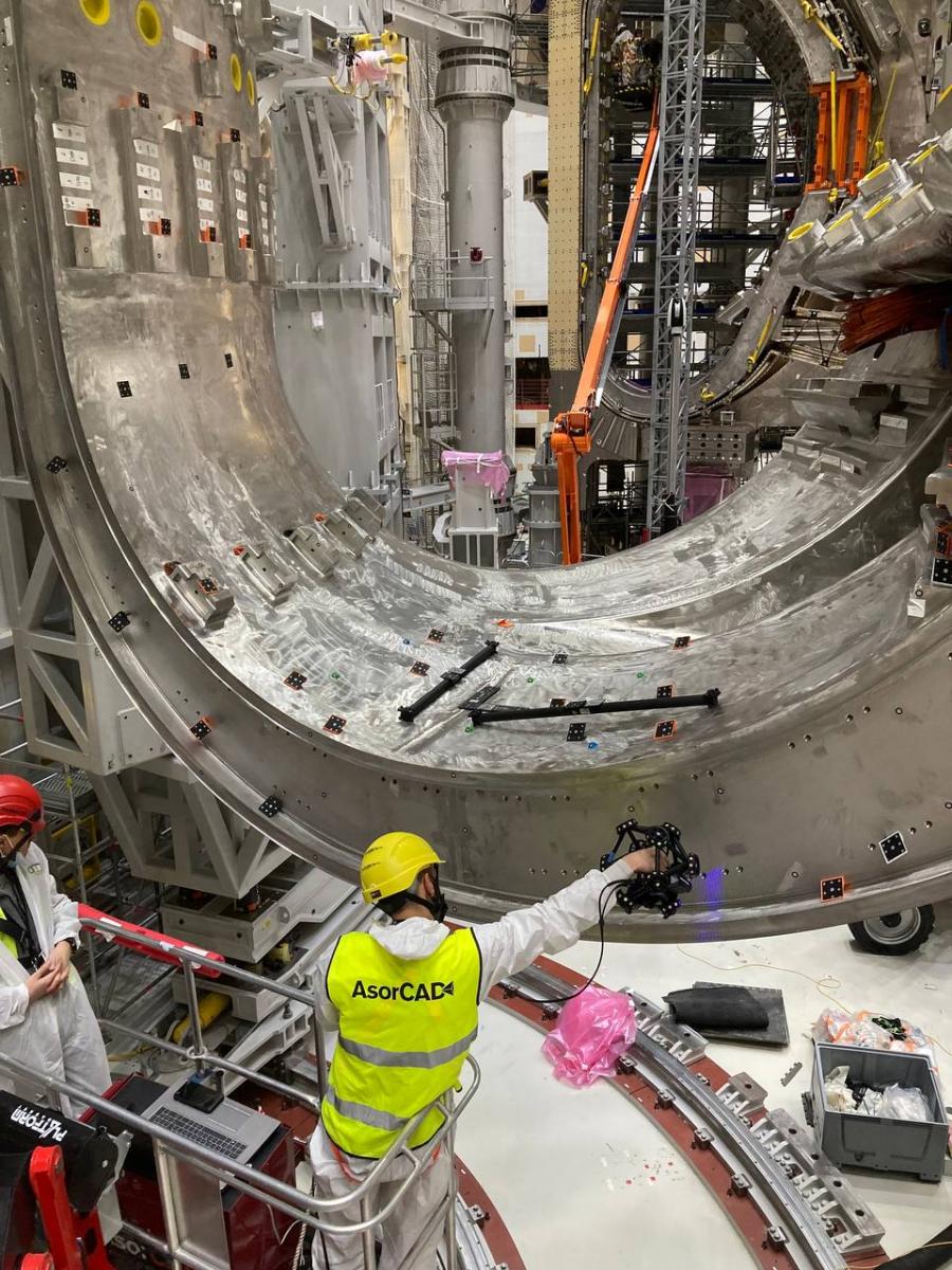 Two engineers wearing safety vests and hard hats inside a boom lift using the MetraCAN 3D scanner to measure a complex part of a tokamak.